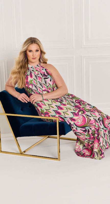 Γυναίκα φορώντας χρωματιστό halter dress από την κολεξιόν Ulla Popken, καθιστή σε navy blue πολυθρόνα